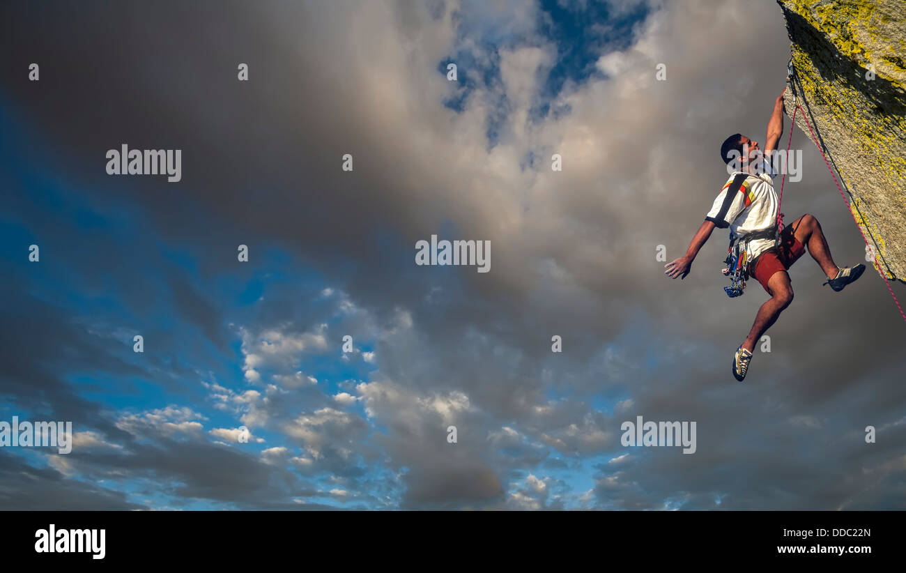 Scalatore di lotte per il vertice di un impegnativo pinnacle come nuvole temporalesche costruire dietro di lui. Foto Stock