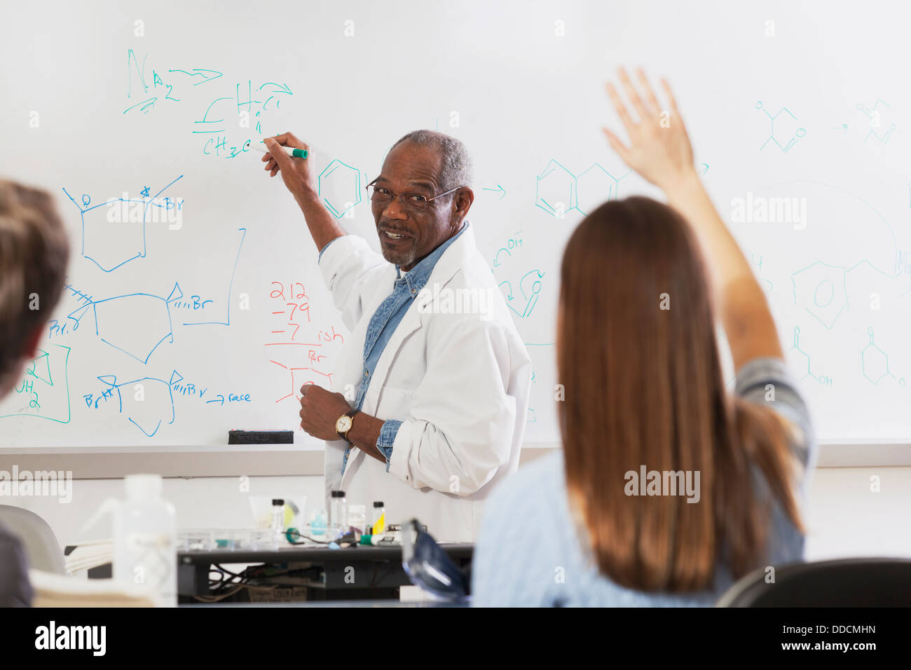 Studente alzando la mano in una classe di ingegneria mentre è professore alla lavagna a scrivere di legami chimici Foto Stock