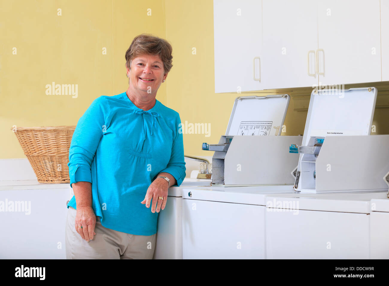 Senior donna in piedi in una sala lavanderia contro una lavatrice commerciale Foto Stock