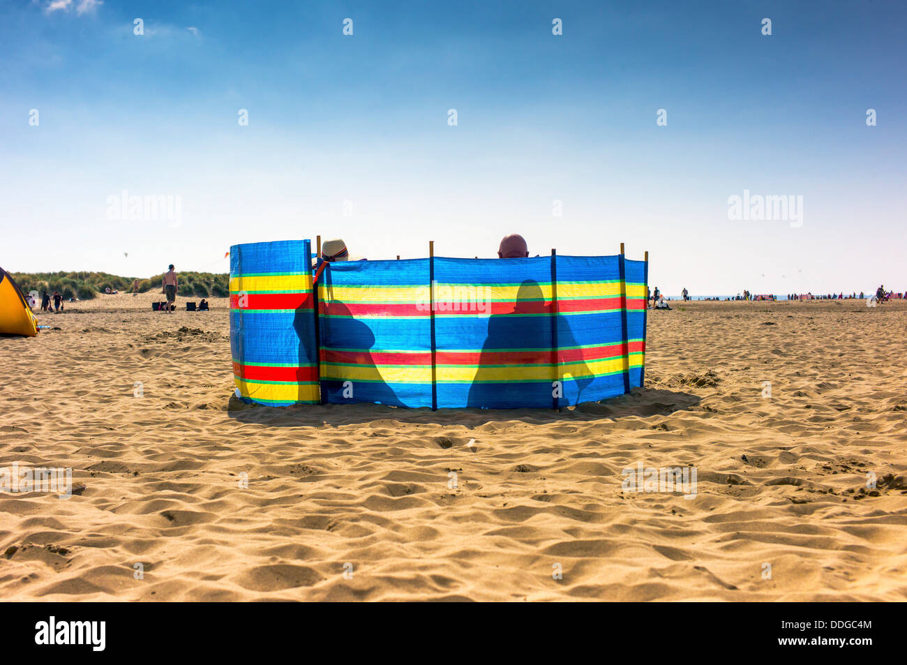 Una coppia matura sit profilarsi dietro un frangivento su una spiaggia, il retro del suo cappello e la sua testa calva visibile sopra la parte superiore, Foto Stock