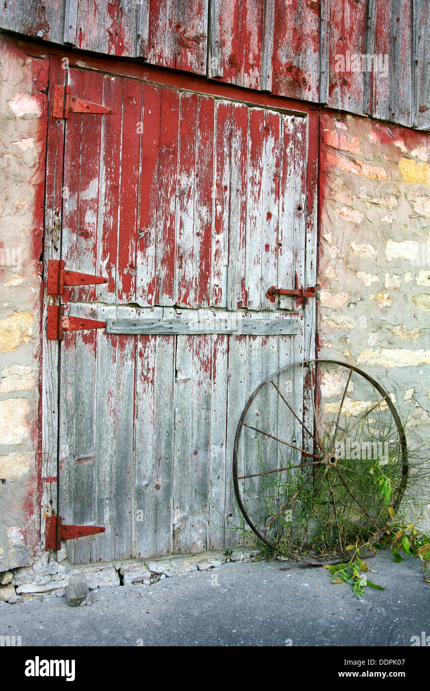 Un rustico vecchio fienile porta con peeling vernice rossa, mura di pietra e un arrugginito antichi ruota del carro Foto Stock