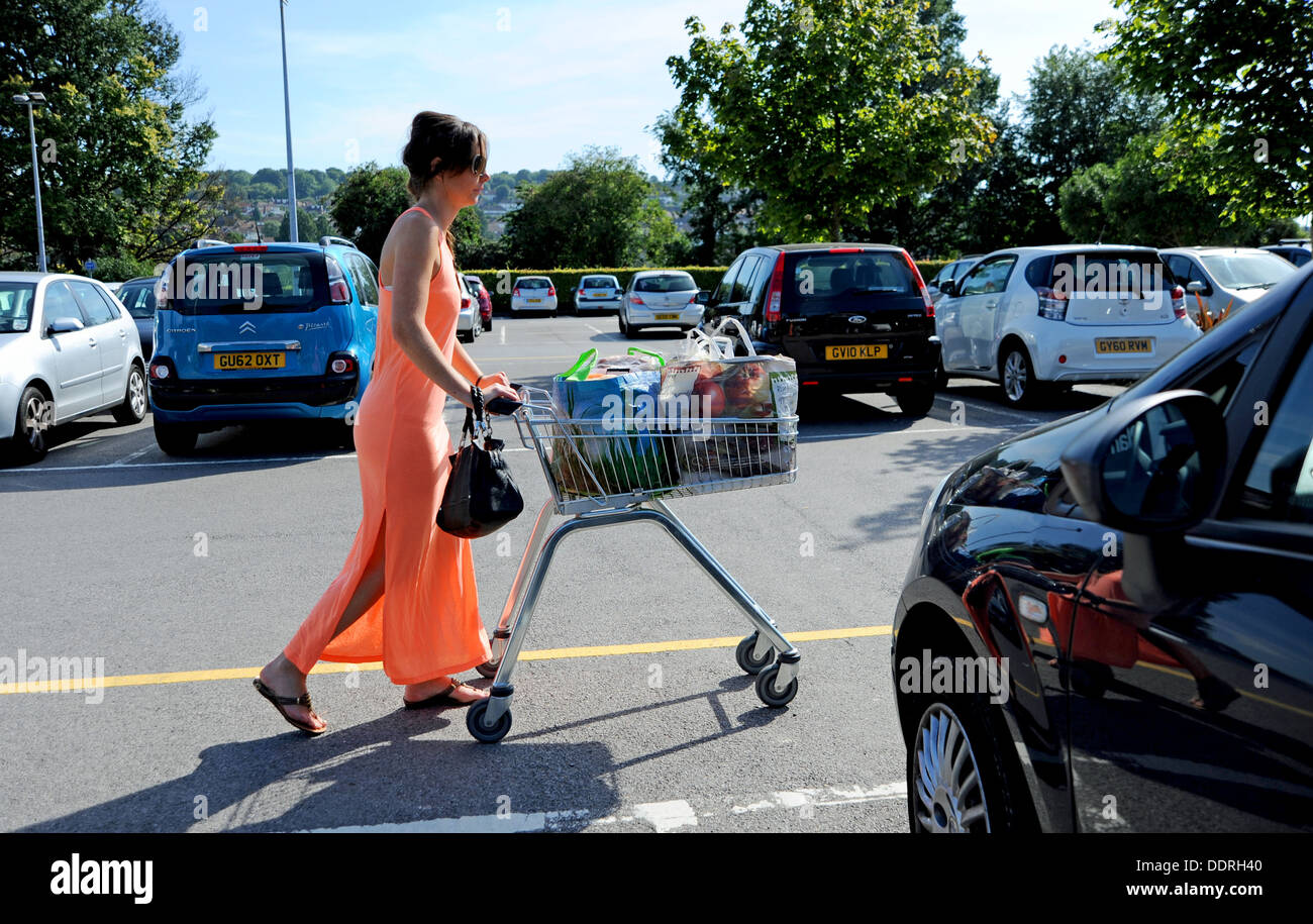 Giovane donna 20s indossando di colore arancione stile maxi abiti estivi spingendo supermercato carrello della spesa nel parco auto Foto Stock