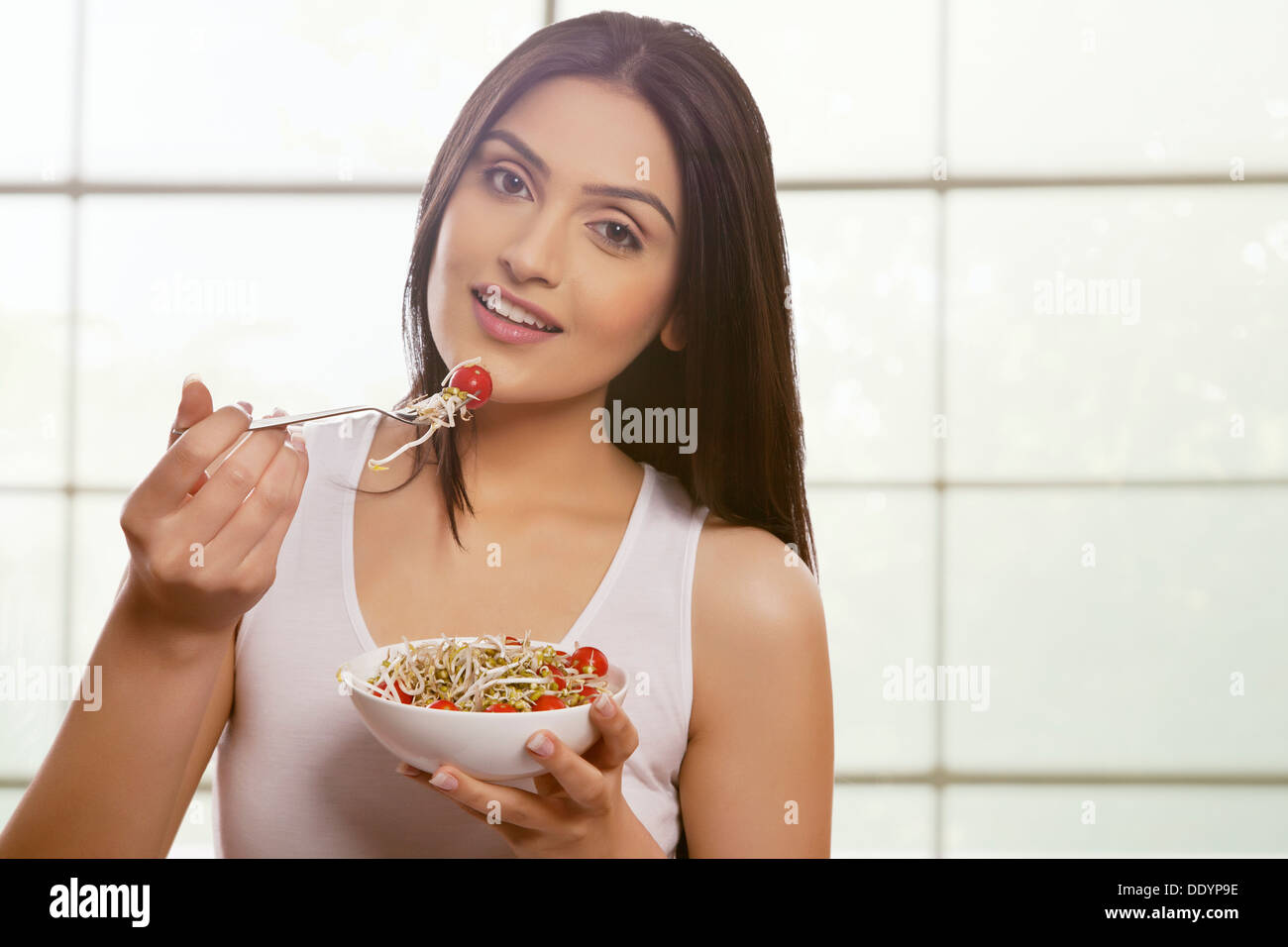 Giovane donna di mangiare i germogli di soia e i pomodori ciliegia contro il vetro della finestra Foto Stock