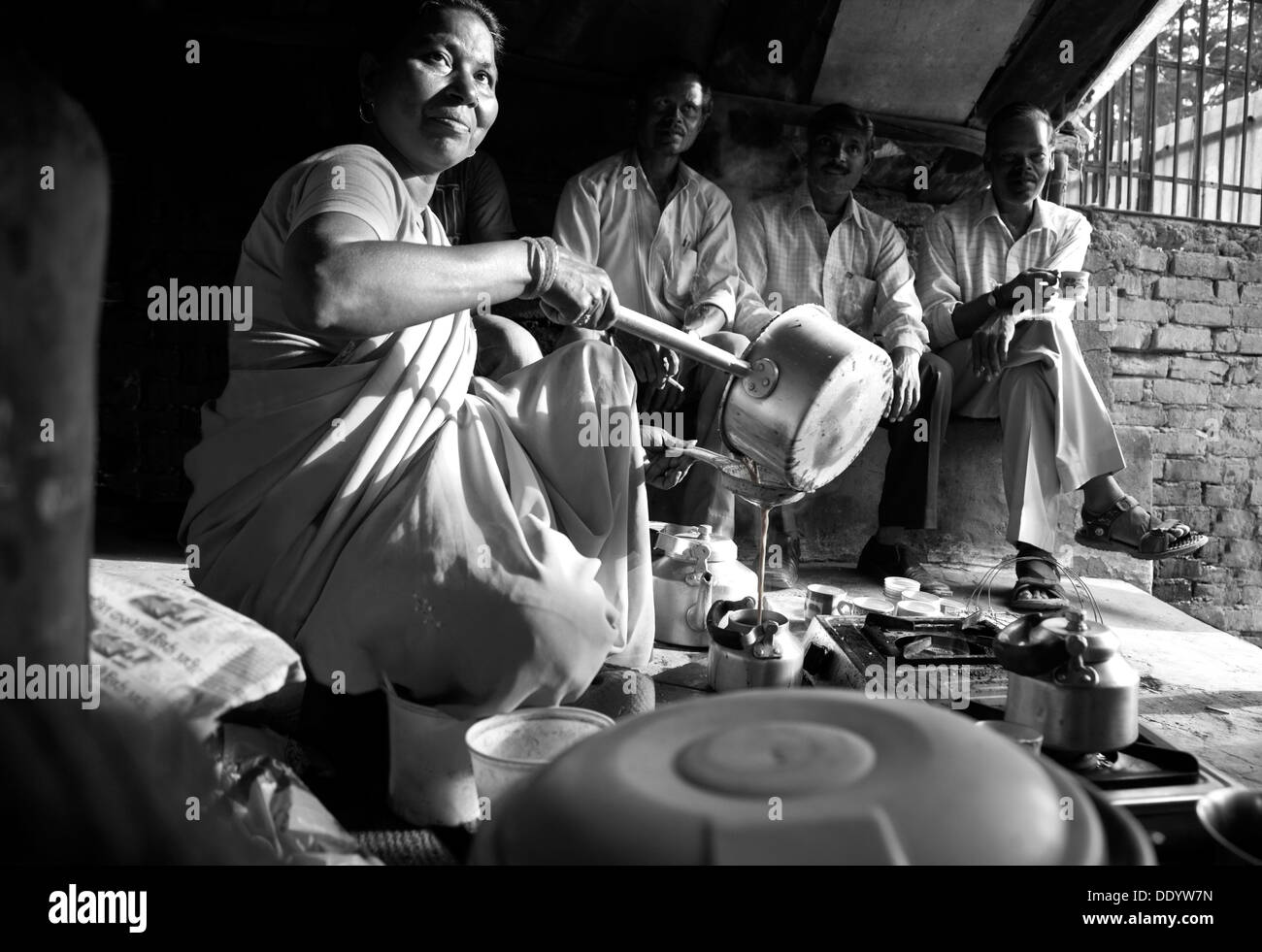 Immagine in bianco e nero di indiani fornitore femmina versando il tè nel bollitore con i clienti seduti in background Foto Stock