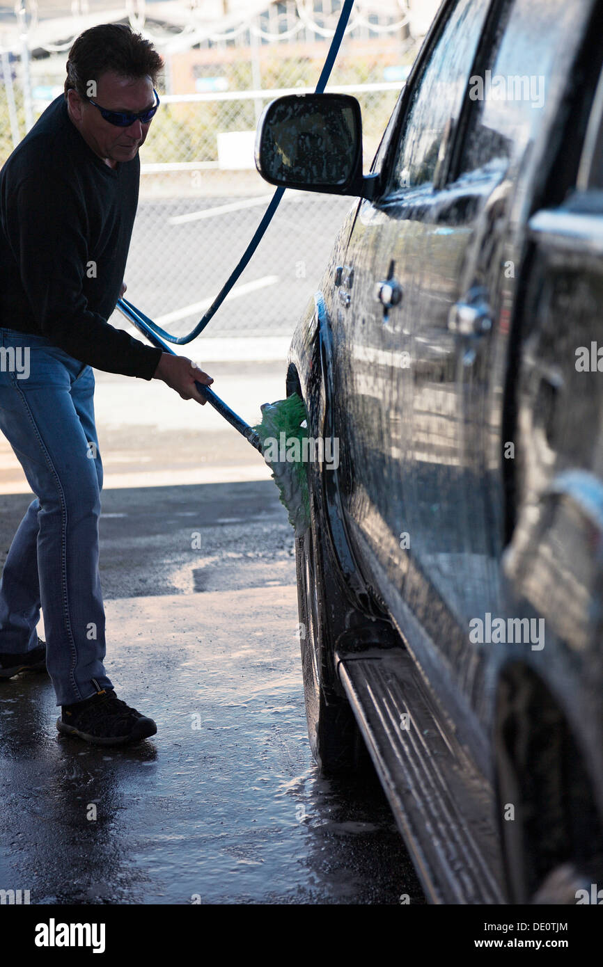 Un uomo con un pennello su un tubo flessibile per pulire il suo veicolo con acqua saponata in corrispondenza di un lavaggio auto. Foto Stock