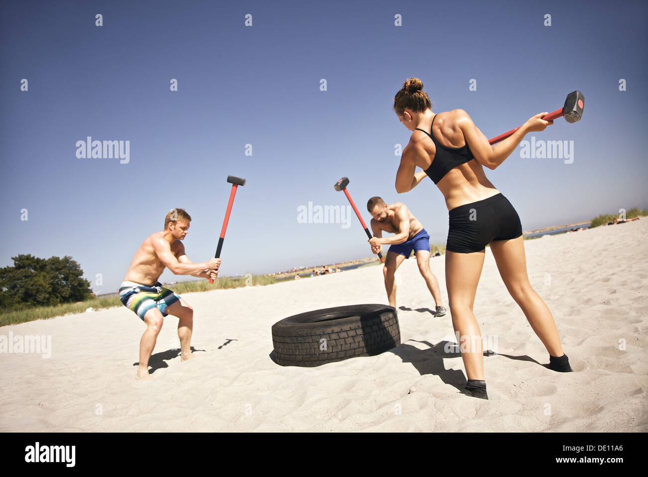 Forte di tre atleti facendo colpo di martello su un carrello pneumatico durante esercizio crossfit fuori sulla spiaggia Foto Stock
