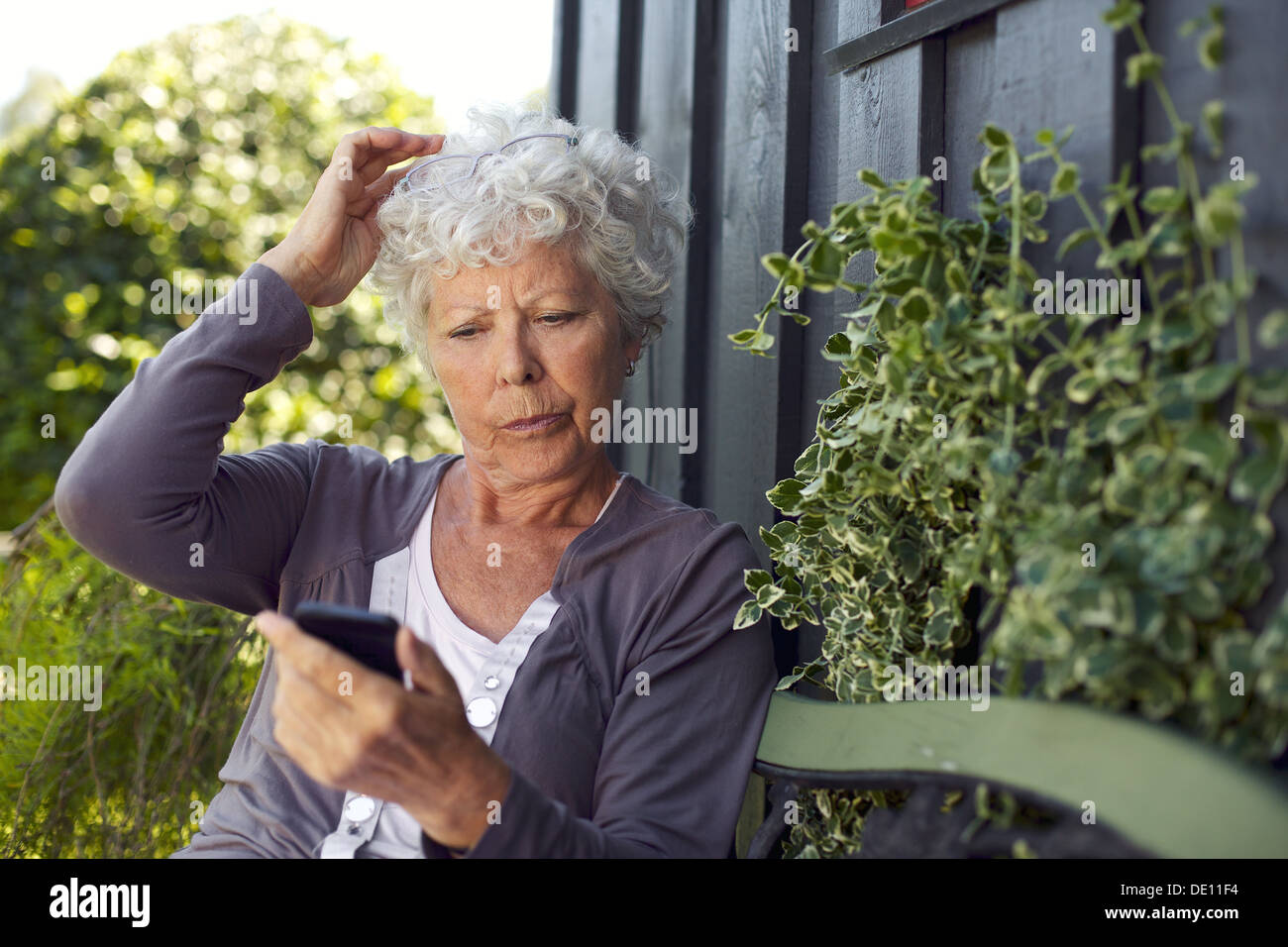Il sambuco donna seduta su una panchina nel suo cortile la lettura di un messaggio di testo sul suo telefono cellulare Foto Stock
