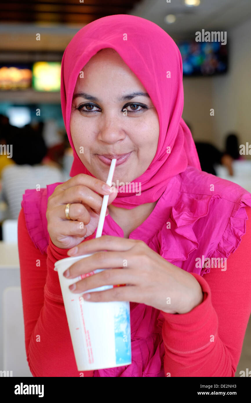 Locale musulmano indonesiano femmina indossando il tradizionale copricapo gustando un drink in un ristorante fast food Foto Stock