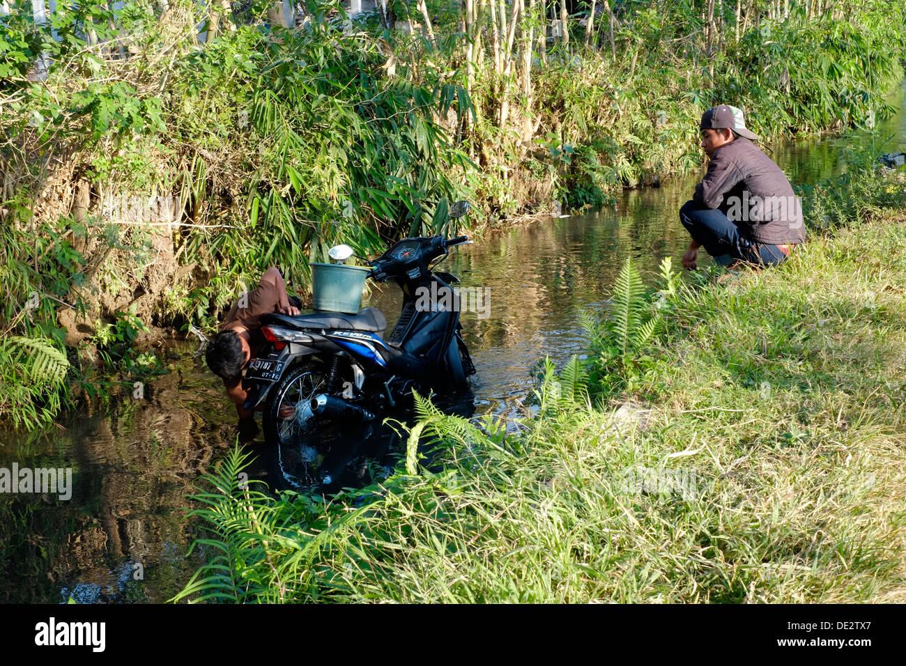 Locali di lavaggio uomo il suo ciclo motore in un piccolo fiume come il suo amico guarda su in un villaggio in java indonesia Foto Stock