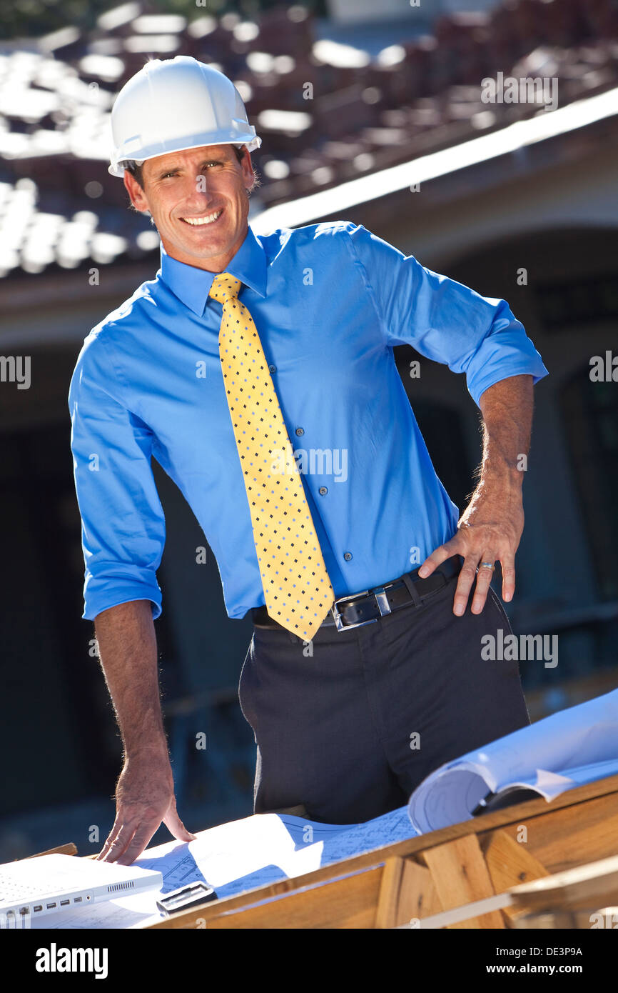 Uomo in elmetto, camicia e cravatta in cantiere con l'architetto piani e un computer portatile. Foto Stock