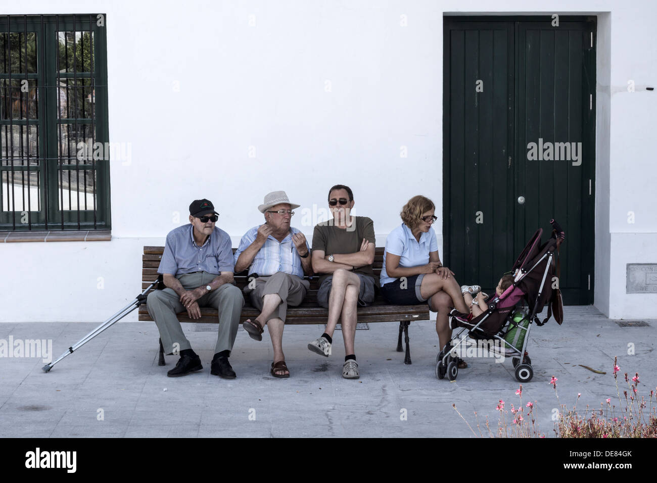 Quattro adulti passare il tempo su un banco a Frigiliana, Spagna, mentre un bambino piccolo guarda dal suo passeggino. Foto Stock