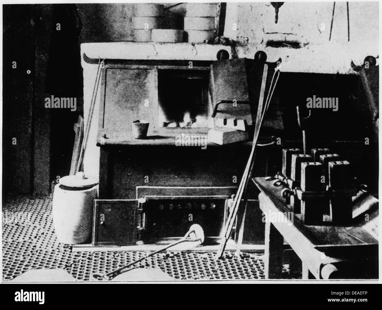 Questa immagine mostra la vista frontale di un forno fusorio con vari strumenti visibili, tra cui un crogiolo, una tazza per immersione, un agitatore, pinze, stampi per lingotti e barre metalliche. La foto evidenzia i processi industriali di lavorazione dei metalli e di produzione. Foto Stock
