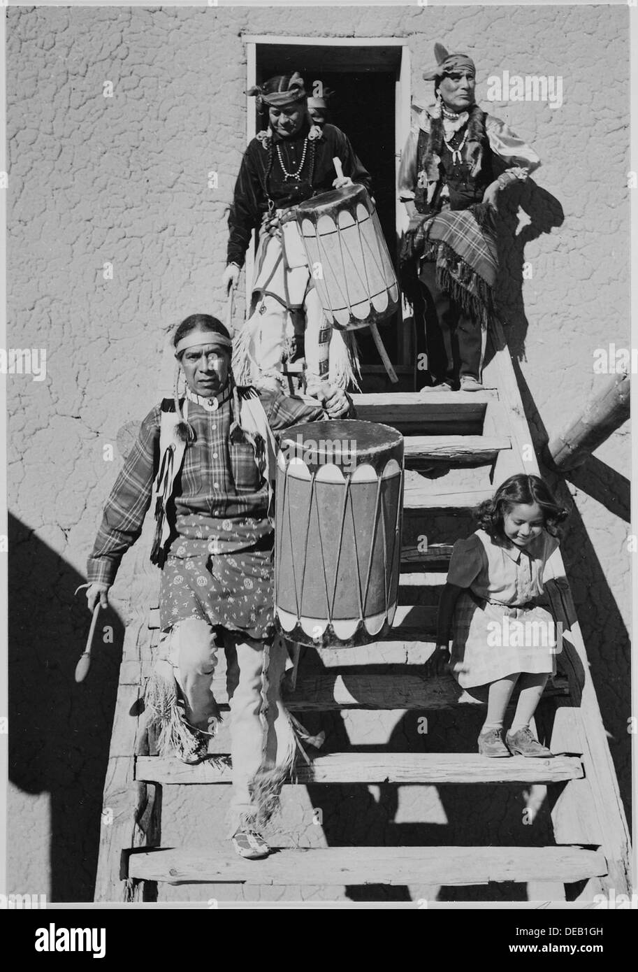 Due indios scendendo le scale in legno, portando i tamburi, un altro Indiano e il bambino vicino alla danza, San Ildefonso Pueblo, Ne 519978 Foto Stock