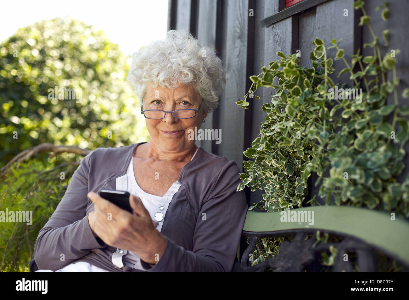 Senior donna utilizzando un telefono cellulare mentre è seduto su un banco di lavoro nel suo giardino nel cortile Foto Stock