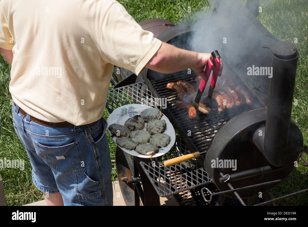 Un uomo nel suo fine 40s Barbecue hamburger e brats su un fumatore/grill nel suo cortile. Wichita, Kansas, Stati Uniti d'America. Foto Stock