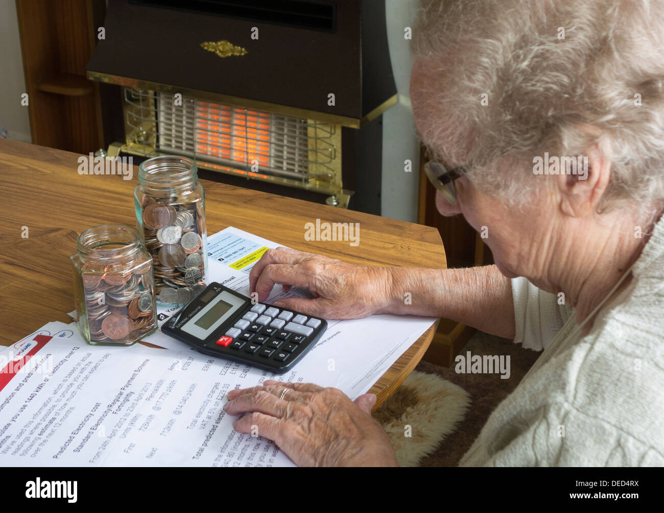 Signora anziana negli anni Novanta che guarda gas, elettricità, bollette con calcolatrice e vasi di monete sul tavolo. Incendio di gas in background Foto Stock