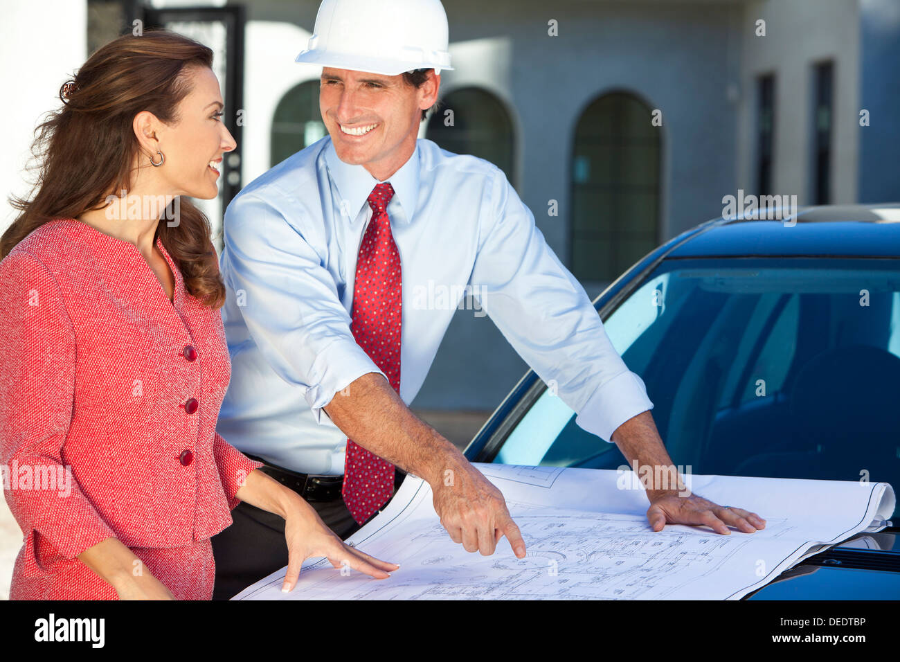 Uomo in elmetto, camicia e cravatta sul sito di costruzione con la donna, guardando l'architetto blu piani di stampa. Foto Stock