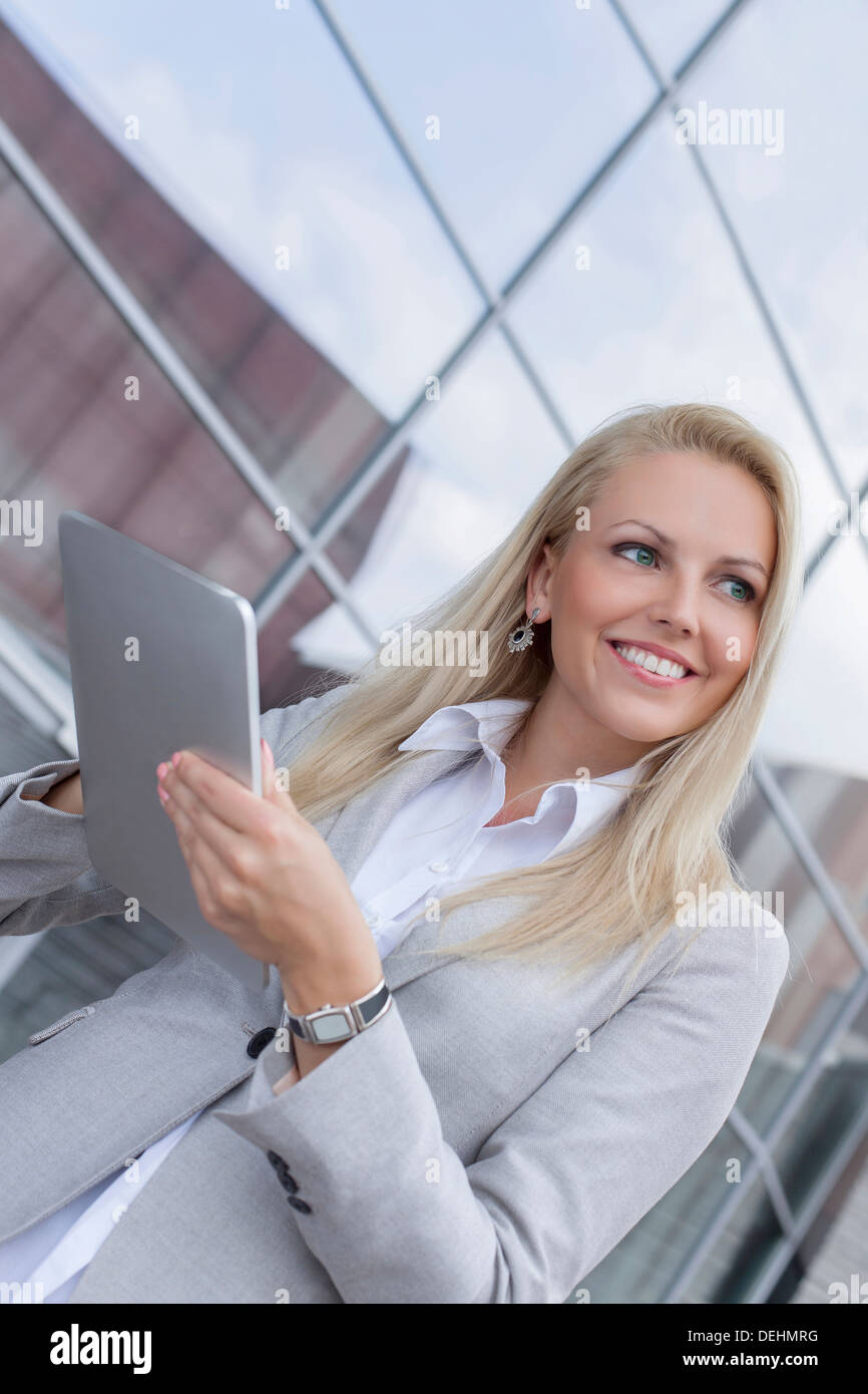 Felice imprenditrice guardando lontano tenendo digitale compressa contro l edificio per uffici Foto Stock