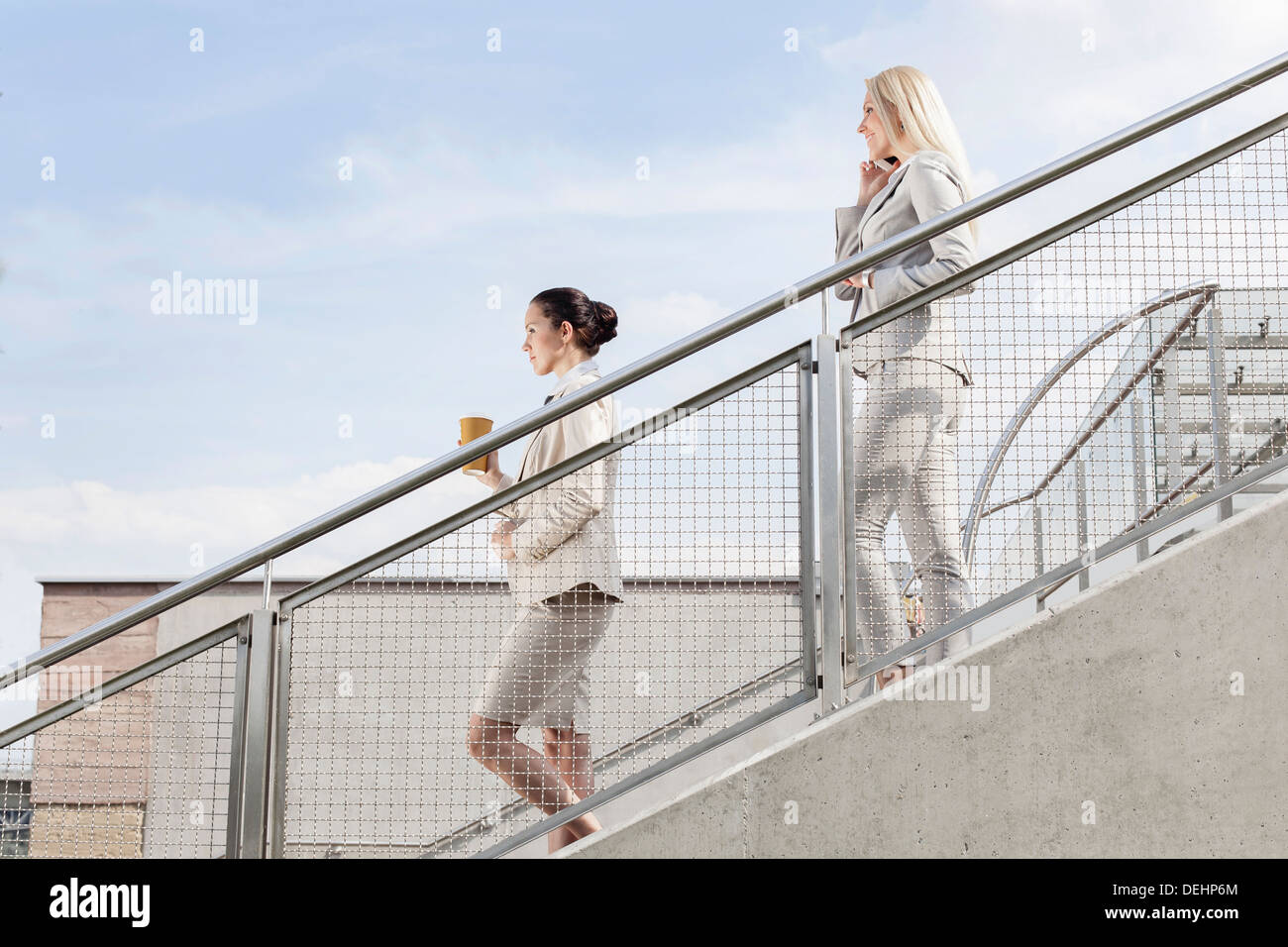 Profilo shot imprenditrici holding tazza monouso telefono cellulare mentre si sposta giù per le scale insieme contro sky Foto Stock