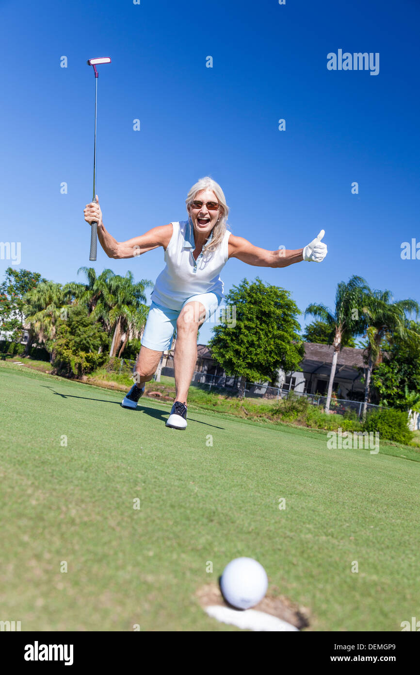 Felice riuscita donna senior giocando a golf putting una pallina da golf in un foro ricavato su di una verde e celebra il suo successo Foto Stock