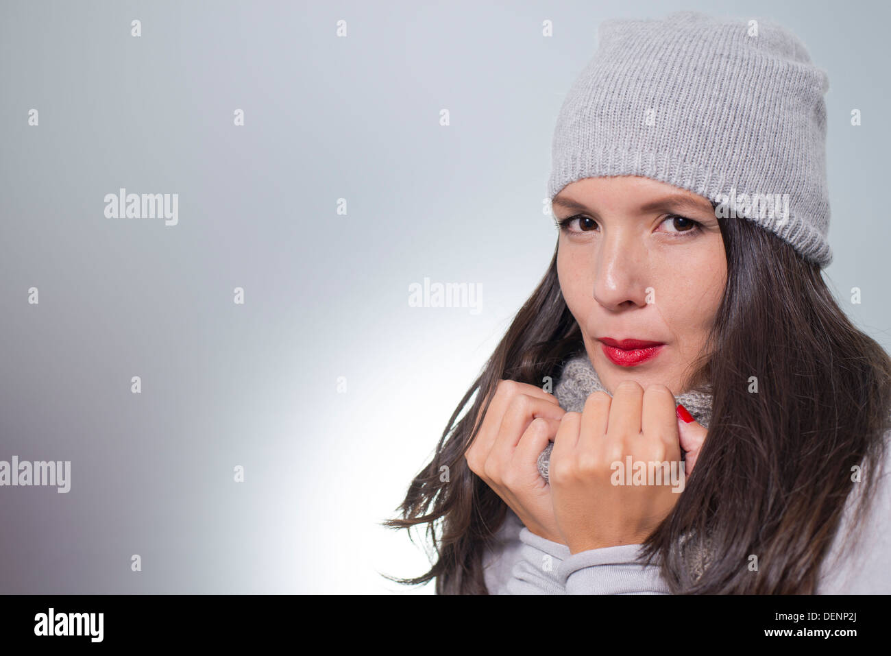 Piuttosto giovane donna in inverno fashion cuddling giù all'interno di lei di lana grigio in maglia jersey, sciarpa e cappello in un freddo giorno Foto Stock