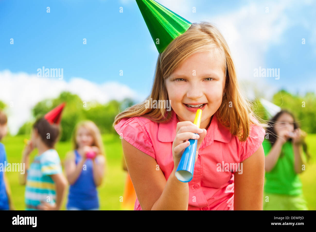 Ritratto di felice ragazza con noisemaker fischio su una festa di compleanno che indossa il cappuccio con amici in piedi sullo sfondo Foto Stock