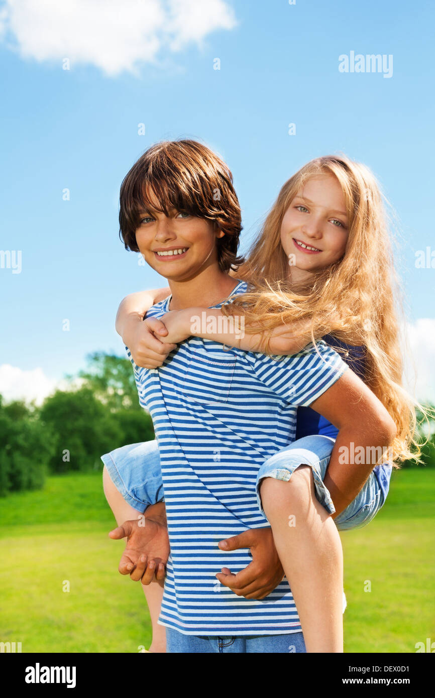 Giovane bella kids, ragazzo portano la ragazza sulla sua schiena, sia felice e sorridente, nel parco sulla soleggiata giornata estiva Foto Stock