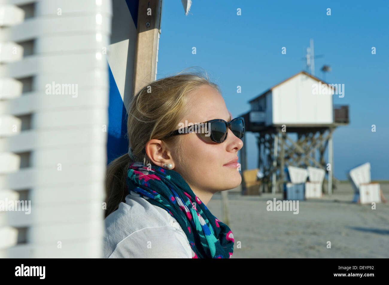 Giovane donna che indossa gli occhiali da sole sulla spiaggia di fronte a una casa su palafitte, St Peter-Ording, Schleswig-Holstein, Germania Foto Stock