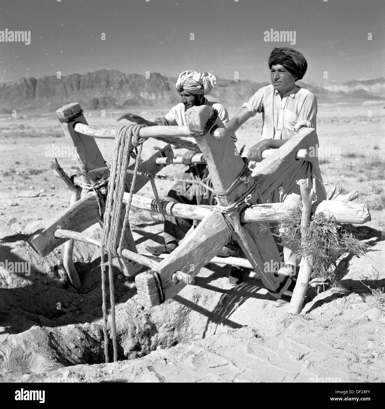 Immagine storica dagli anni '50 di J Allan Cash di due uomini afghani locali che girano le maniglie di legno di un rachet di legno sopra un buco d'acqua o pozzo fuori nel mezzo di una zona rurale del deserto dell'Afghanistan. Foto Stock