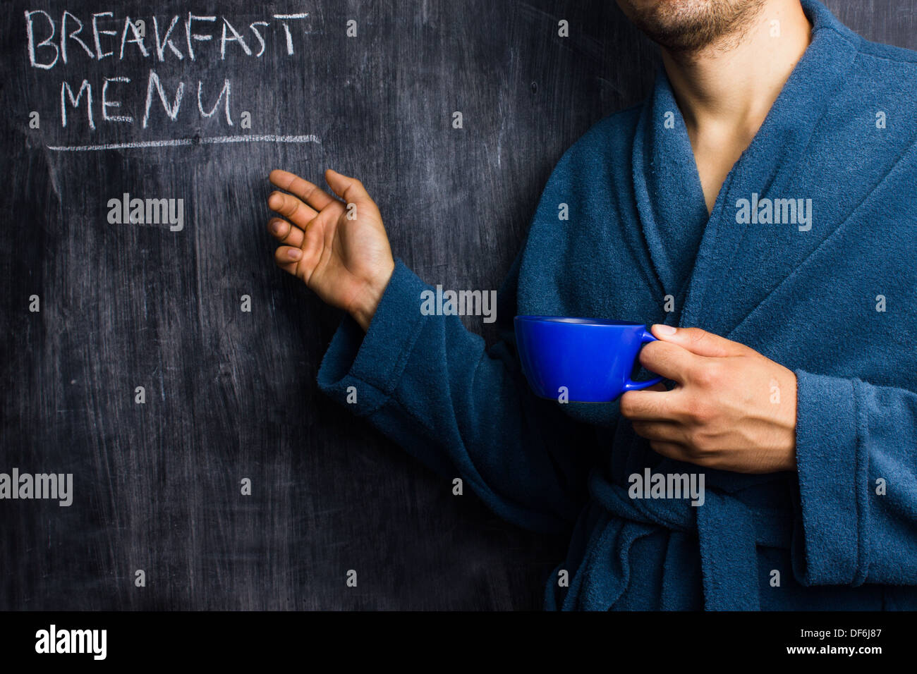 Giovane uomo in accappatoio è in piedi da lavagna con una tazza di caffè e puntando verso il menu della prima colazione Foto Stock