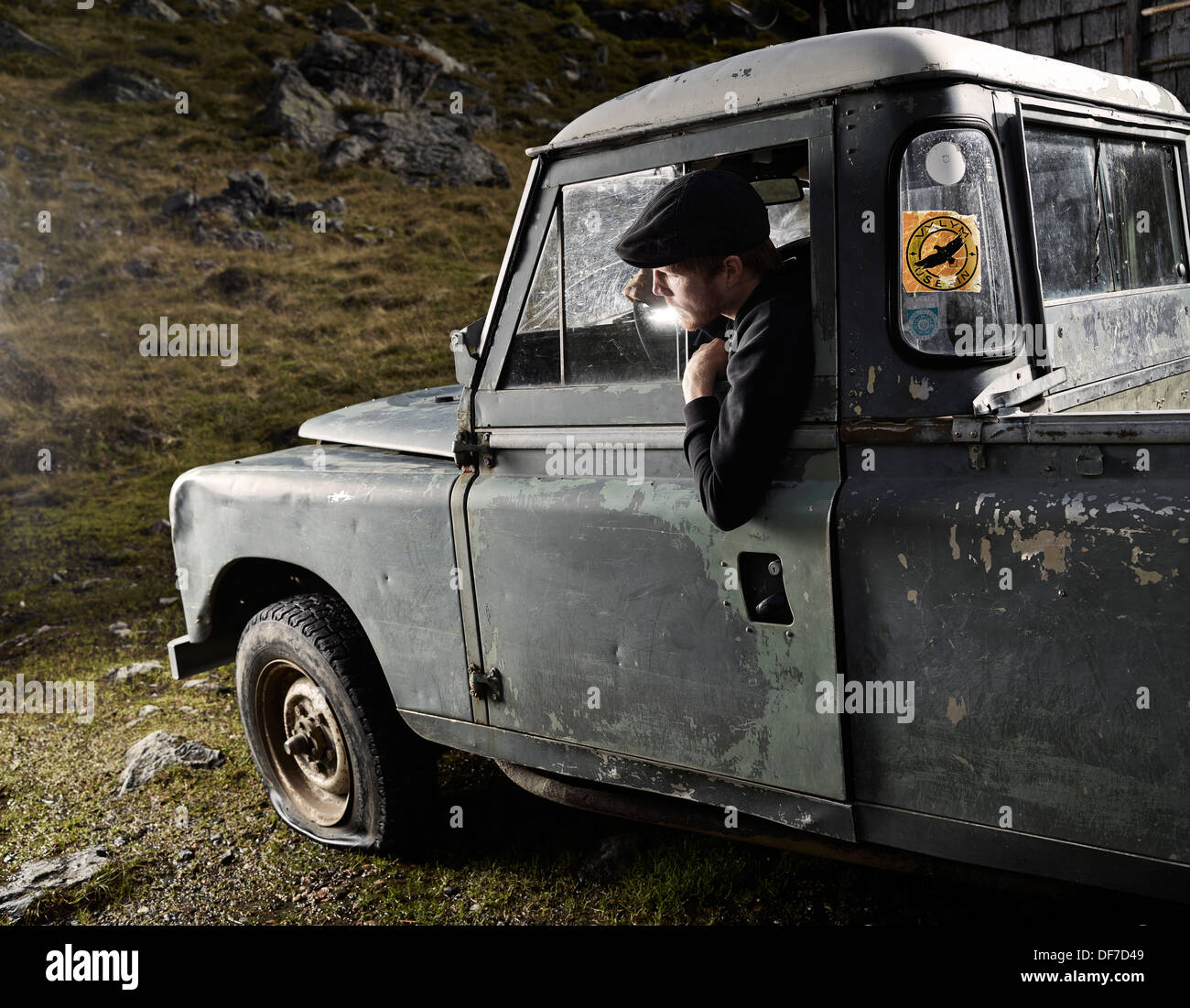 Uomo con un cappello seduto nel sedile del conducente di una vettura vecchia notando un appartamento di pneumatico anteriore sinistro, Inneralpbach, Tirolo, Austria Foto Stock