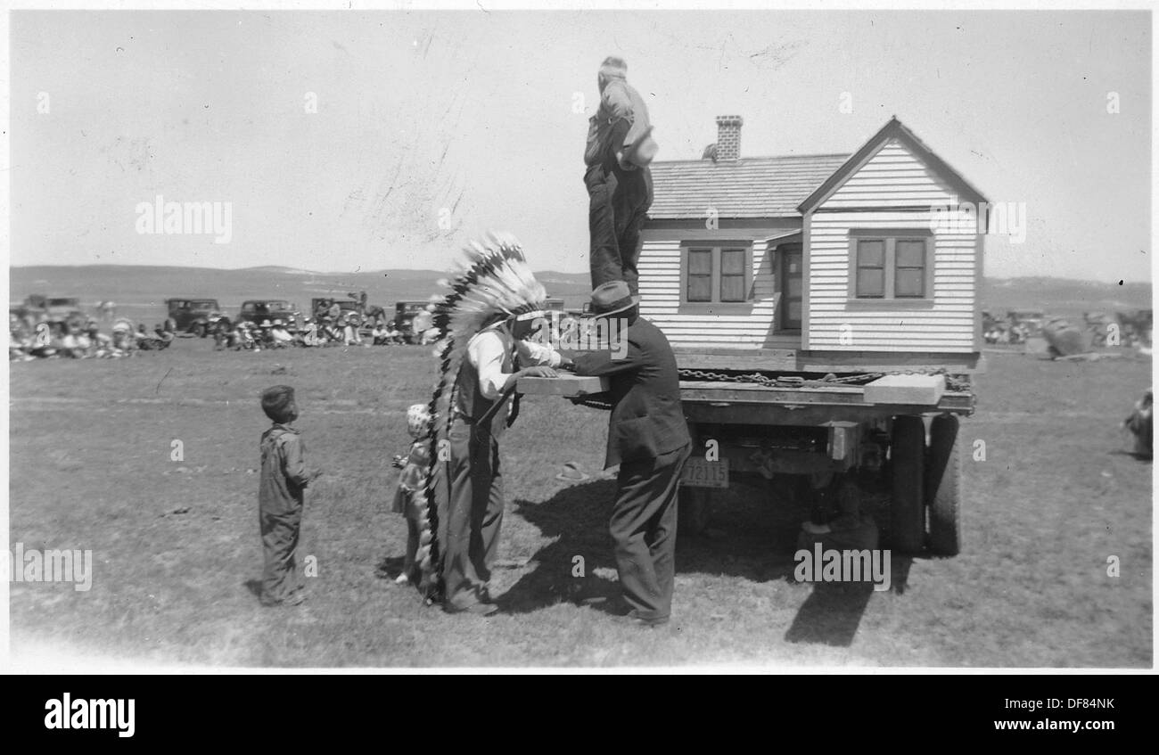 Questa immagine mostra un venditore e un individuo nativo americano in piedi accanto a una casa modello costruita in scala, esposta sul retro di un camion nella città di Oglala. La scena fa parte di uno sforzo per promuovere l'edilizia abitativa moderna nelle comunità dei nativi americani, riflettendo gli sforzi di sviluppo del XX secolo volti a migliorare le condizioni di vita nelle riserve. Foto Stock