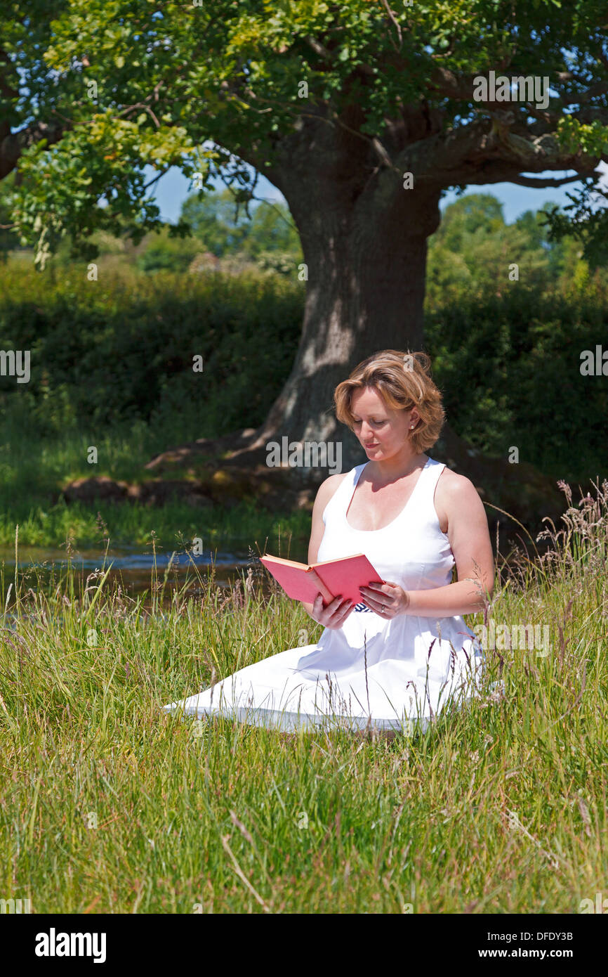 Una donna in abito bianco, seduti in un prato oltre ad una corrente di lettura di un libro su una luminosa giornata d'estate. Foto Stock