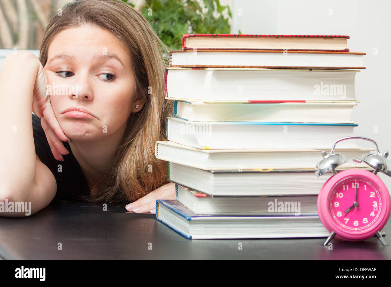 Una giovane donna sguardi al grande pila di libri per lo studio dell'esame. Tempo zecche da ricordandole il tempo limitato a disposizione per preparare. Foto Stock