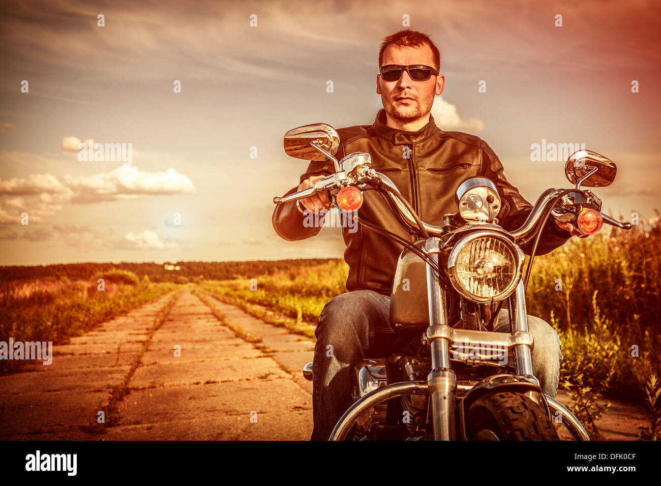 Biker uomo che indossa una giacca di pelle e occhiali da sole seduto sulla sua moto guardando il tramonto. Foto Stock Biker uomo che indossa una giacca di pelle e occhiali da sole seduto sulla sua moto guardando il tramonto. Foto Stock