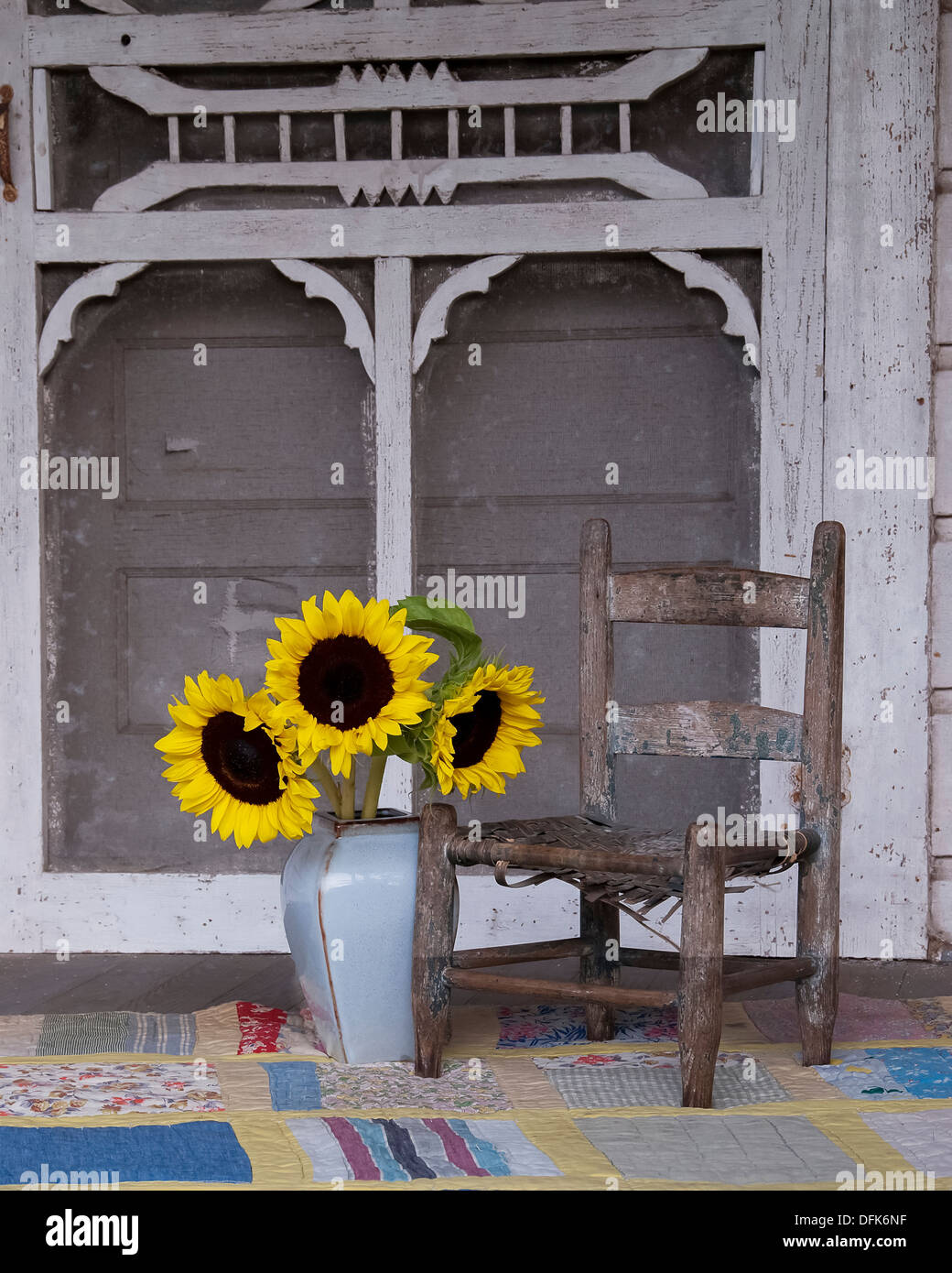 Una vecchia sedia, Vaso con girasoli, una trapunta su un portico di ruggine con una porta dello schermo come sfondo. Foto Stock