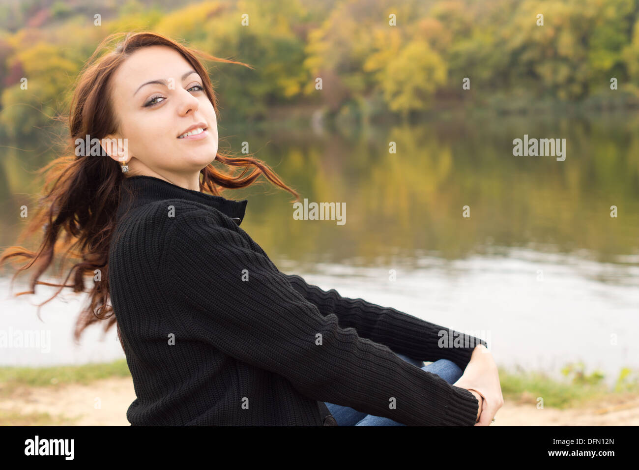 Felice giocoso giovane donna seduta al di sopra di un tranquillo fiume o lago tornando indietro a guardare la fotocamera con i suoi lunghi capelli bruna Foto Stock