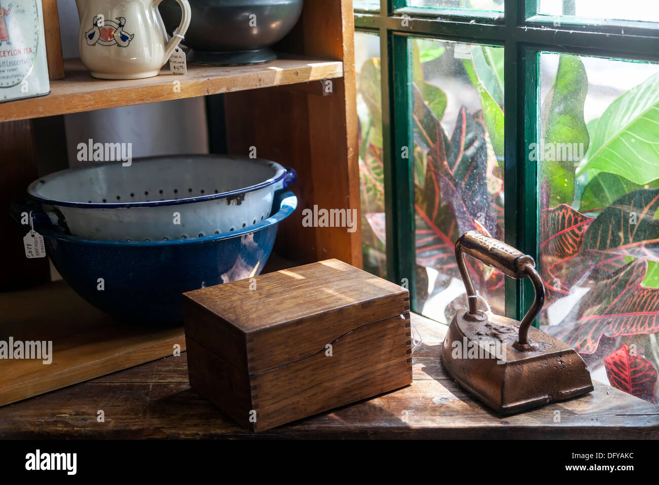 Antique vintage ghisa ferro da stiro, scatola di legno e metallo smaltato scolapasta e ciotola su scaffalature di legno da una finestra. Foto Stock