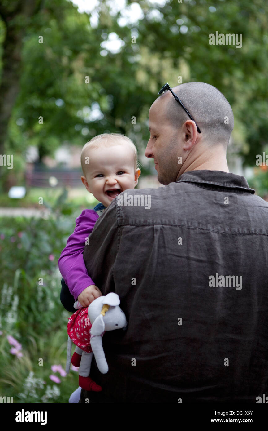 Baby girl ridendo e aggraffatura giocattolo morbido, guardando oltre il padre della spalla. Foto Stock
