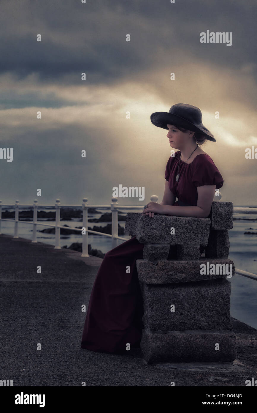Una ragazza triste con un cappello nero seduto su una panchina di pietra al mare Foto Stock