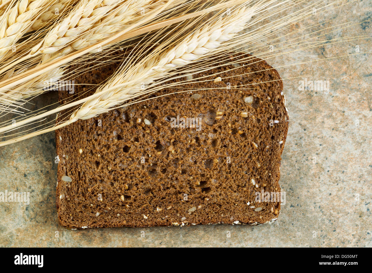 Foto orizzontale di una singola fetta di dolce, scuro tutto il pane di grano essiccato con gli steli di grano sulla pietra naturale bancone Foto Stock