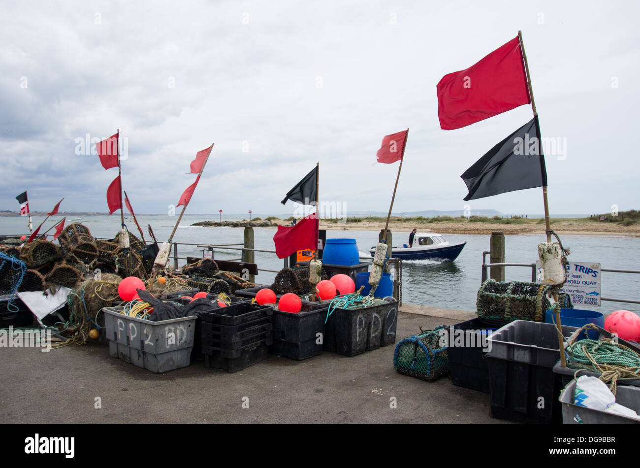 Una scena in un villaggio di pescatori di Mudeford con attrezzi da pesca e bandiere rosse su una riva del mare e barche che passano in una giornata di mare. Foto Stock