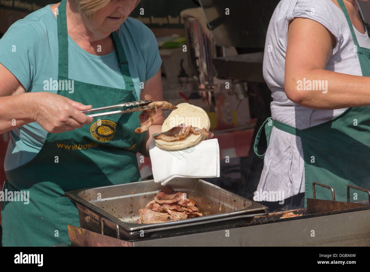 Stallholder mettendo le fette spesse di succosa, grasso di pancetta a caldo su di un grande panino pane alla Bowland stallo a Bolton Food Festival 2013. Foto Stock