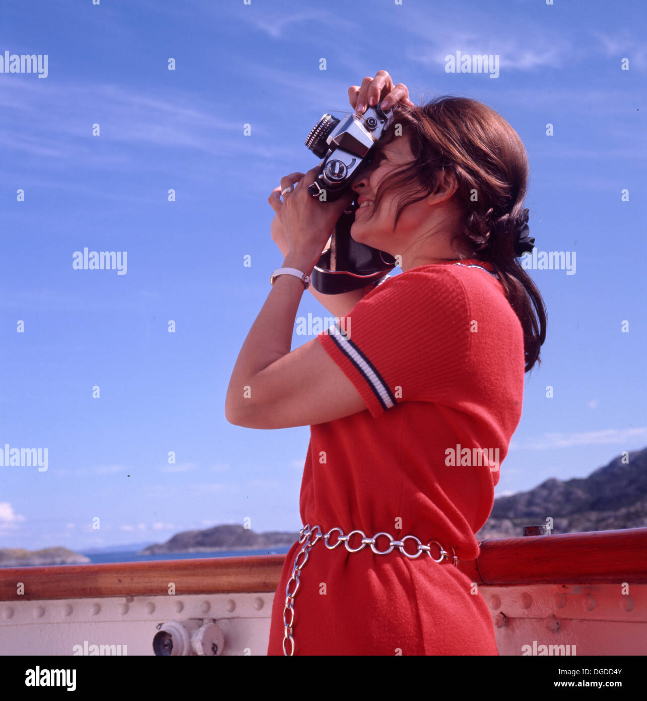 anni '1970, turista donna in barca in abito rosso che utilizza la luce brillante del cielo, tiene una fotocamera da 35 mm all'occhio e si concentra per scattare una foto. Foto Stock