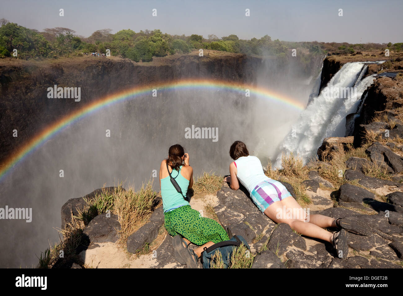 Victoria Falls Zambia; Due turisti donne che guardano oltre il bordo alle Cascate Vittoria con arcobaleno, lato Zambia, Livingstone Island, Zambia Africa Foto Stock