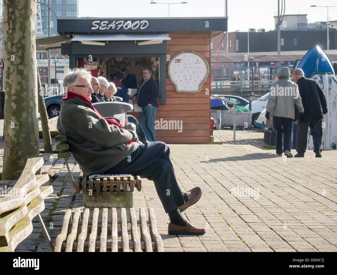 Un uomo che indossa abiti caldi solo seduta su una panchina sul disco di Portsmouth con una pressione di stallo di frutti di mare in background Foto Stock