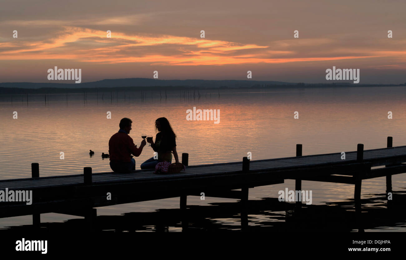 Silhouette di un paio nel crepuscolo seduto su di un molo in legno sul lago la tostatura con vino rosso bicchieri, Steinhude am Meer Foto Stock
