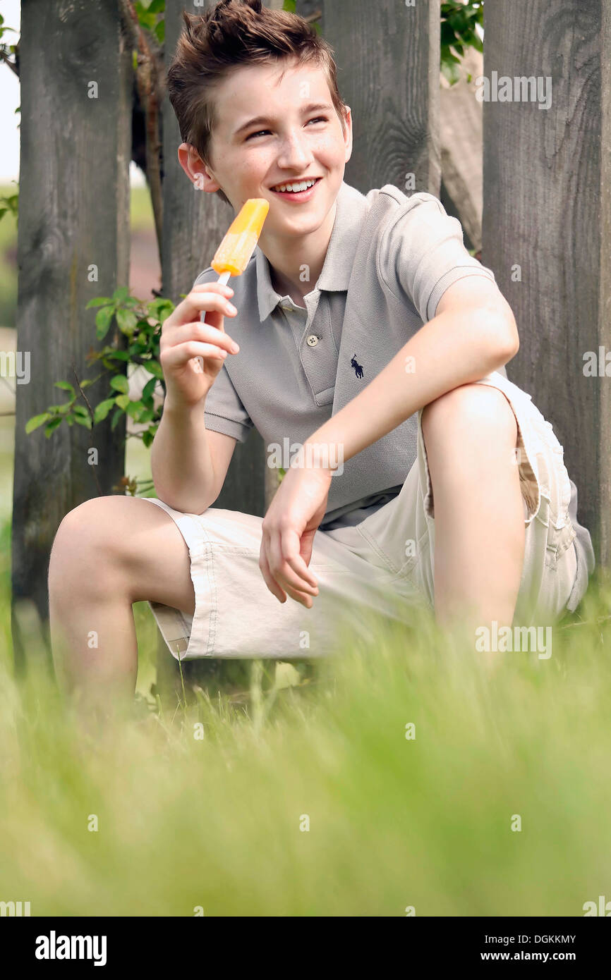 Ragazzo (12-13) seduto in erba e mangiare il gelato Foto Stock