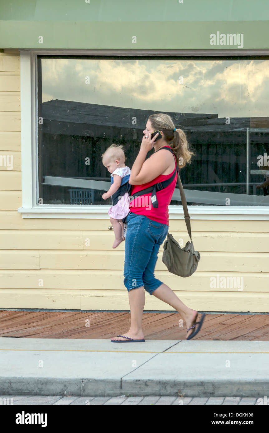 Giovane donna madre indossa maglietta rossa e pantaloni capri che porta il bambino neonato mentre parlano al telefono cellulare. Foto Stock