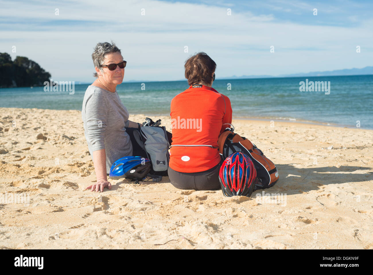 Due donne (età 50+) sulla spiaggia in Nuova Zelanda chiacchierando e guardando il mare, con ciclo caschi e indossando indumenti attivo Foto Stock